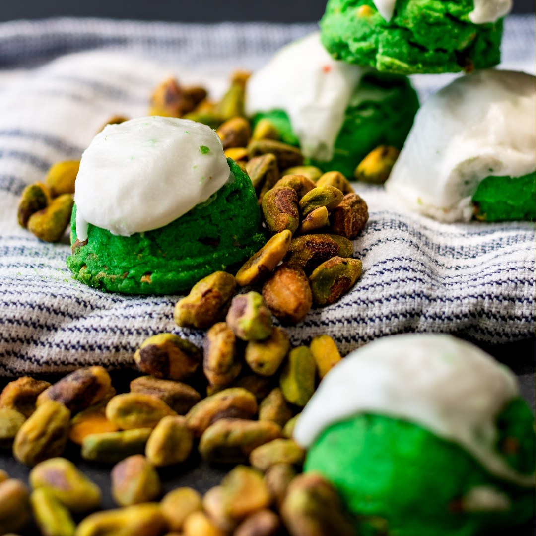 Green pistachio cookies topped with white icing nestled in pistachios on a blue-striped kitchen towel.