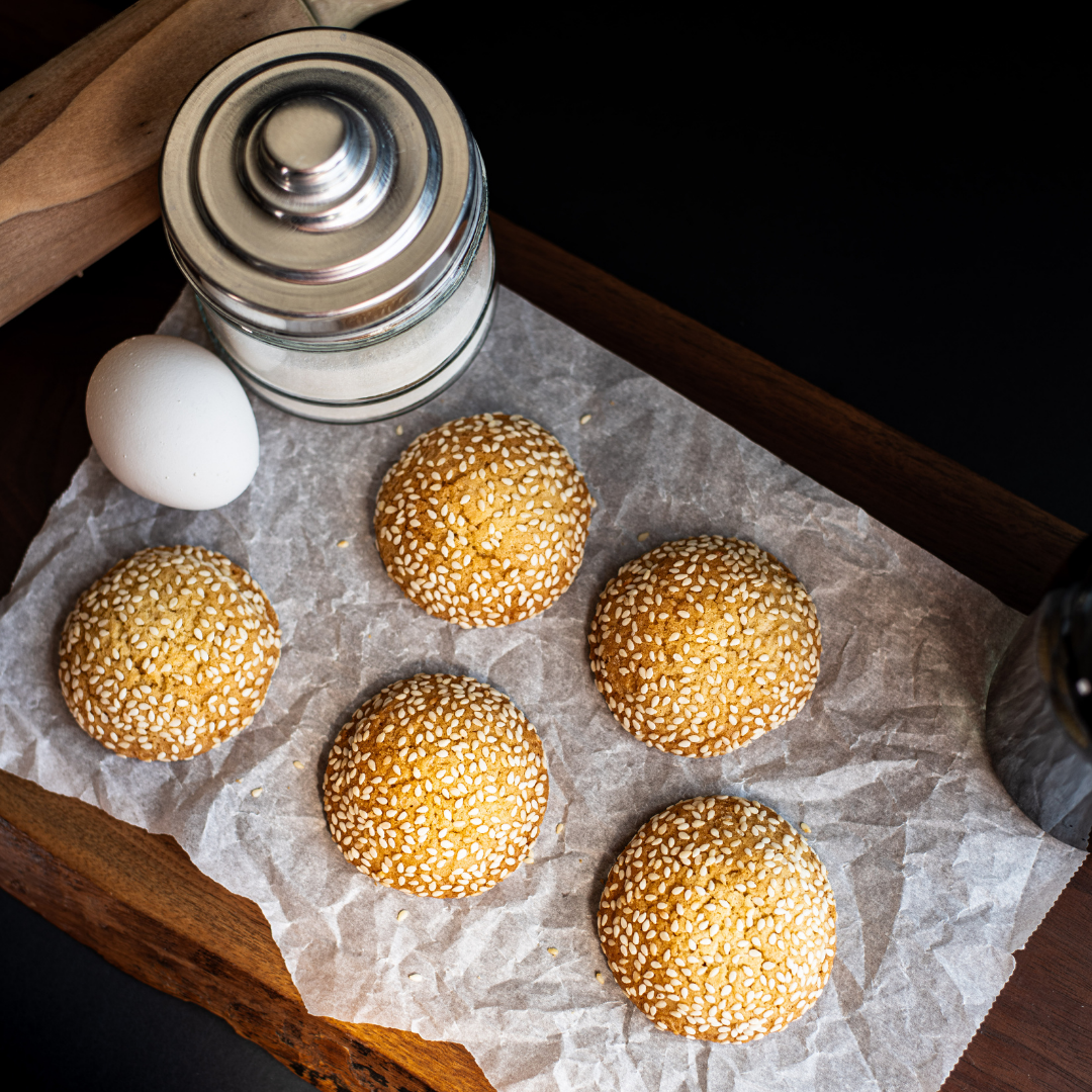 Six sesame cookies arranged on parchment paper with an egg and sugar jar on a dark wood board.