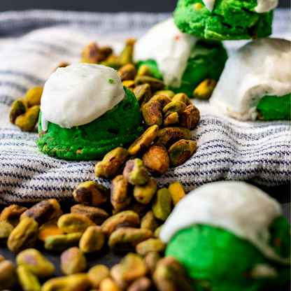 Green pistachio cookies topped with white icing nestled in pistachios on a blue-striped kitchen towel.