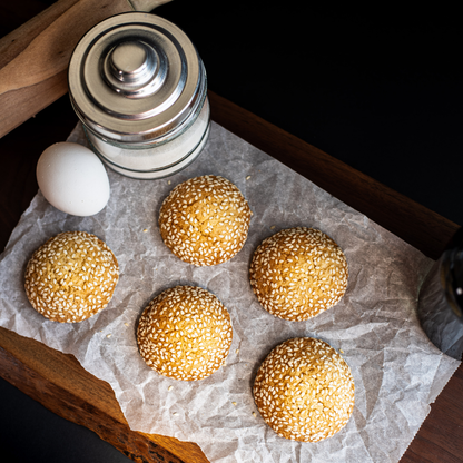 Six sesame cookies arranged on parchment paper with an egg and sugar jar on a dark wood board.